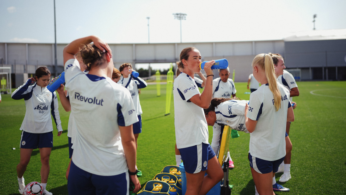 Group of female soccer players wearing Manchester City training gear, drinking water, and relaxing on a field during training.