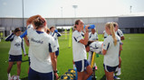 Group of female soccer players wearing Manchester City training gear, drinking water, and relaxing on a field during training.