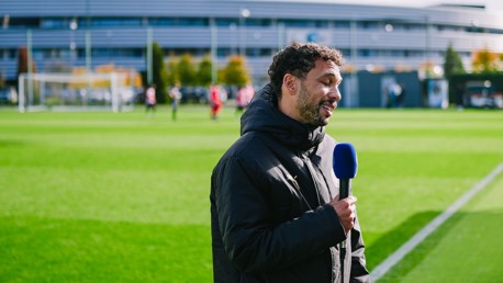 A man in a black jacket holds a microphone with a blue windscreen, speaking near a sports field. The background shows people playing on a green field with a building in the distance.