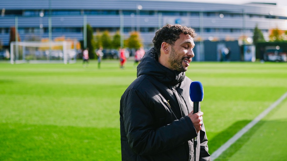 A man in a black jacket holds a microphone with a blue windscreen, speaking near a sports field. The background shows people playing on a green field with a building in the distance.