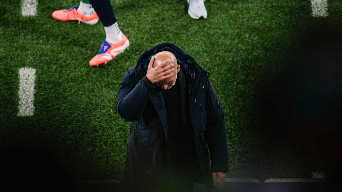 A football coach in a black jacket holds his head with one hand, standing on a green field with players nearby.