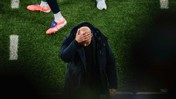 A football coach in a black jacket holds his head with one hand, standing on a green field with players nearby.