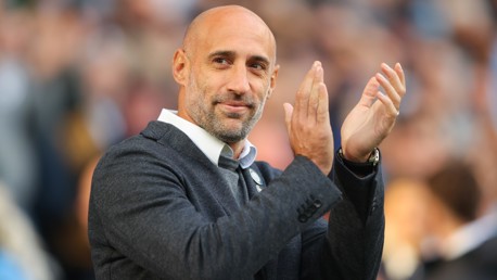 A coach clapping on the sidelines wearing a dark jacket and white shirt, with the background showing a blurred crowd.