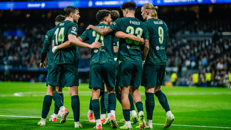 Manchester City players celebrating a goal on the field during a match, wearing their dark green away kit with names 'Mathus Nunes,' 'O'Reilly,' and 'Haaland' visible.