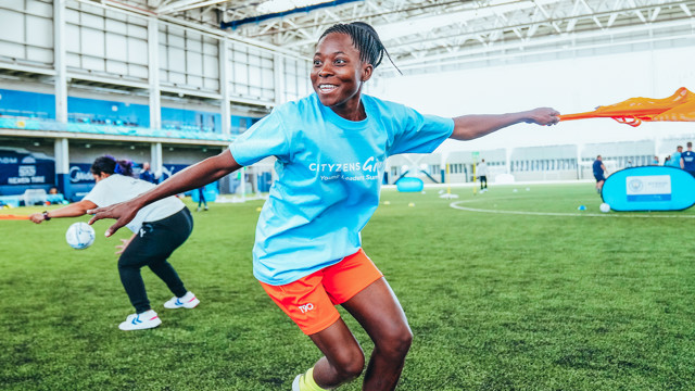 A person in an indoor sports facility wearing a Cityzens Giving shirt and holding a lacrosse stick, participating in a sports activity.