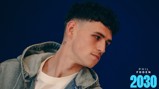 A person with short, curly hair wearing a denim jacket and white shirt, looking sideways against a blue background. Text on the right includes 'Phil Foden 2030.'