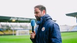 Person holding a microphone with Manchester City logo, wearing a blue jacket at a football stadium.