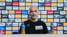 A bald man at a press conference with Manchester City branding behind him, wearing a black shirt marked 'EM' and 'Asahi'. Lucozade bottles are on the table.