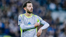 A Manchester City player wearing a grey and neon green kit, adjusting the captain's armband during a Premier League match. The player's face is blurred.
