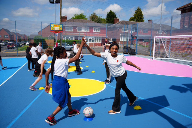 Children wearing white t-shirts engage in a football activity on a colorful outdoor court, high-fiving each other. The event takes place in a neighborhood with houses visible in the background.
