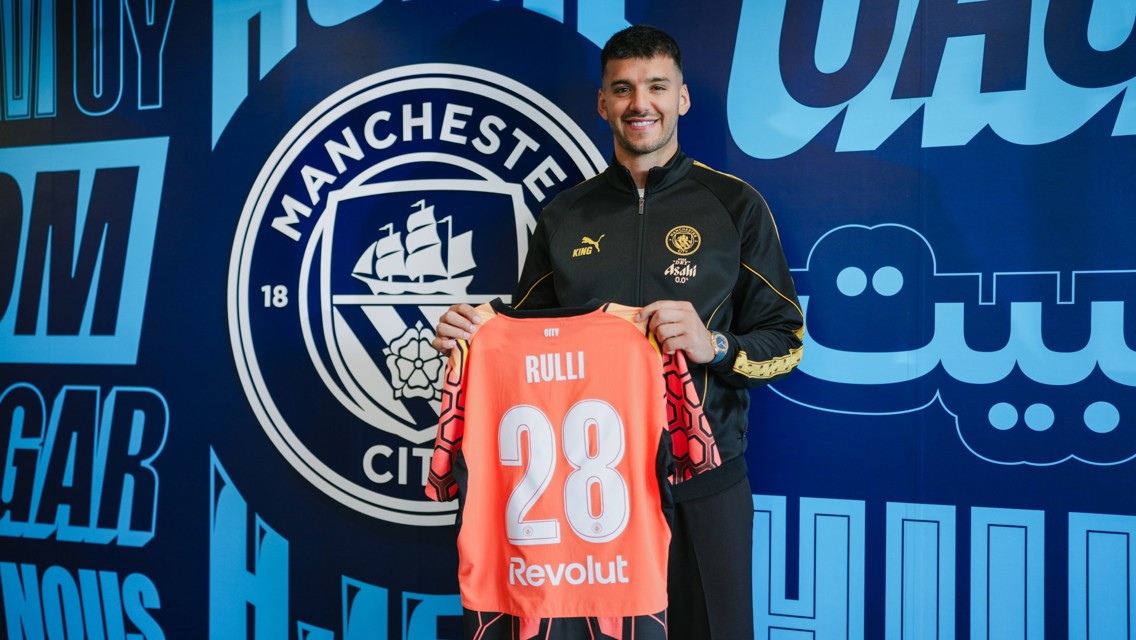 A person holding a Manchester City jersey with the name RULLI and number 28 inside the club's facility with Manchester City logo in the background.