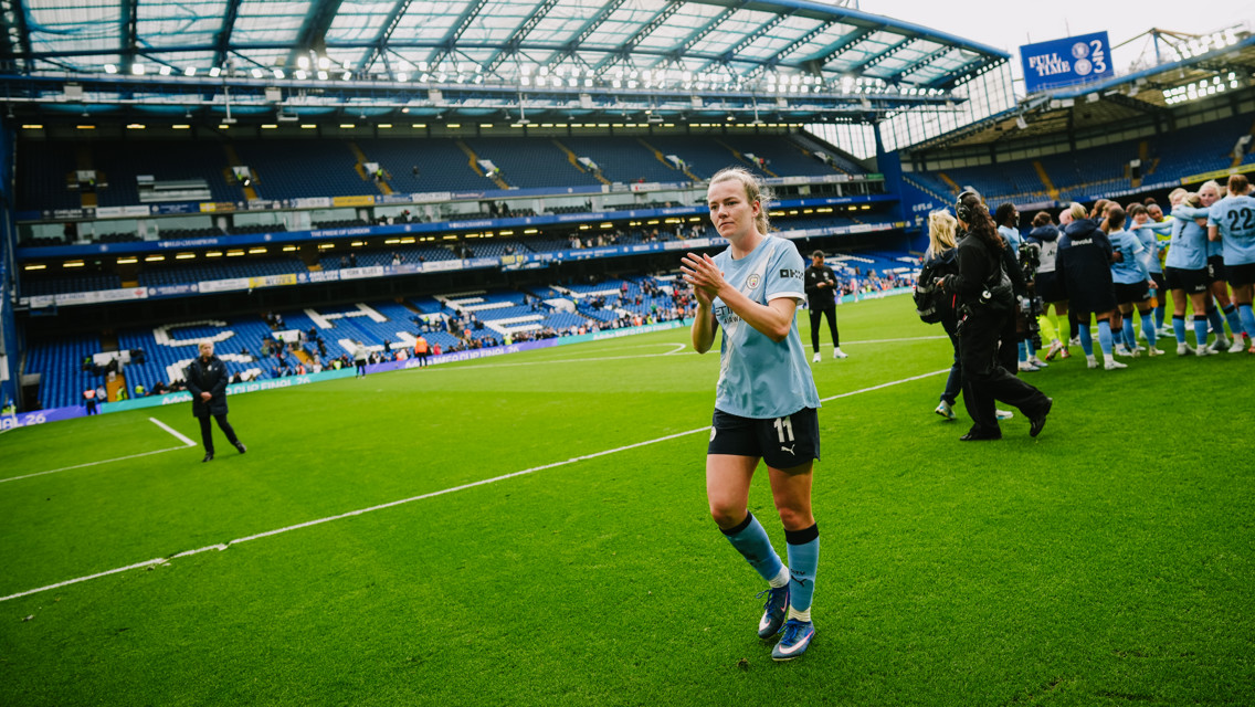 A football player in a light blue jersey claps hands on the pitch, with teammates and camera crew nearby at a stadium.