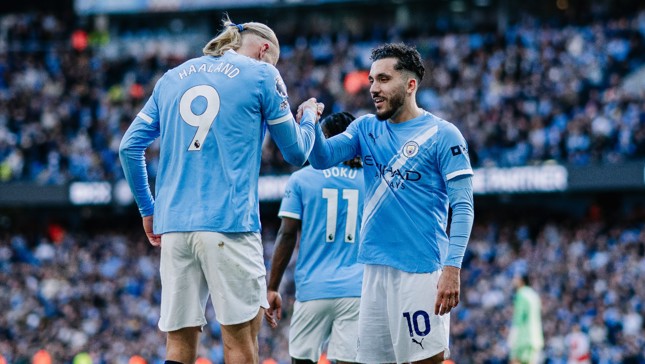 Several football players in Manchester City jerseys are celebrating during a match. The players have numbered jerseys, and the scene is set in a stadium with a crowd in the background.