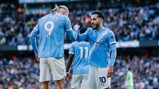Several football players in Manchester City jerseys are celebrating during a match. The players have numbered jerseys, and the scene is set in a stadium with a crowd in the background.