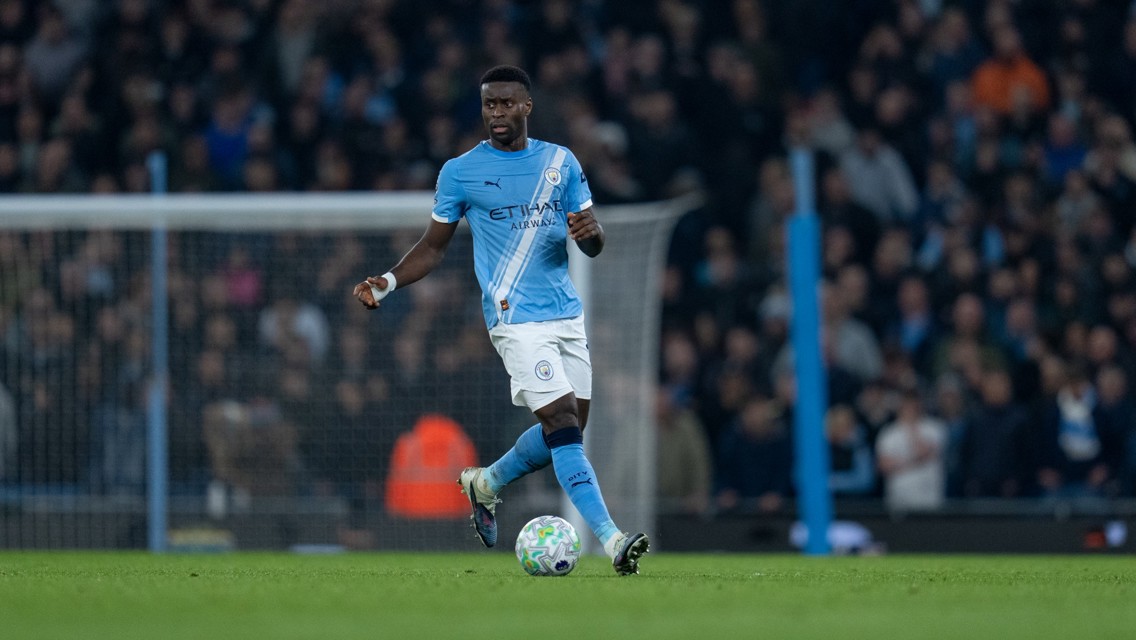 A football player from Manchester City is controlling the ball in a stadium, wearing the team's light blue kit. The background shows a crowd of spectators.