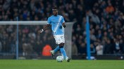 A football player from Manchester City is controlling the ball in a stadium, wearing the team's light blue kit. The background shows a crowd of spectators.