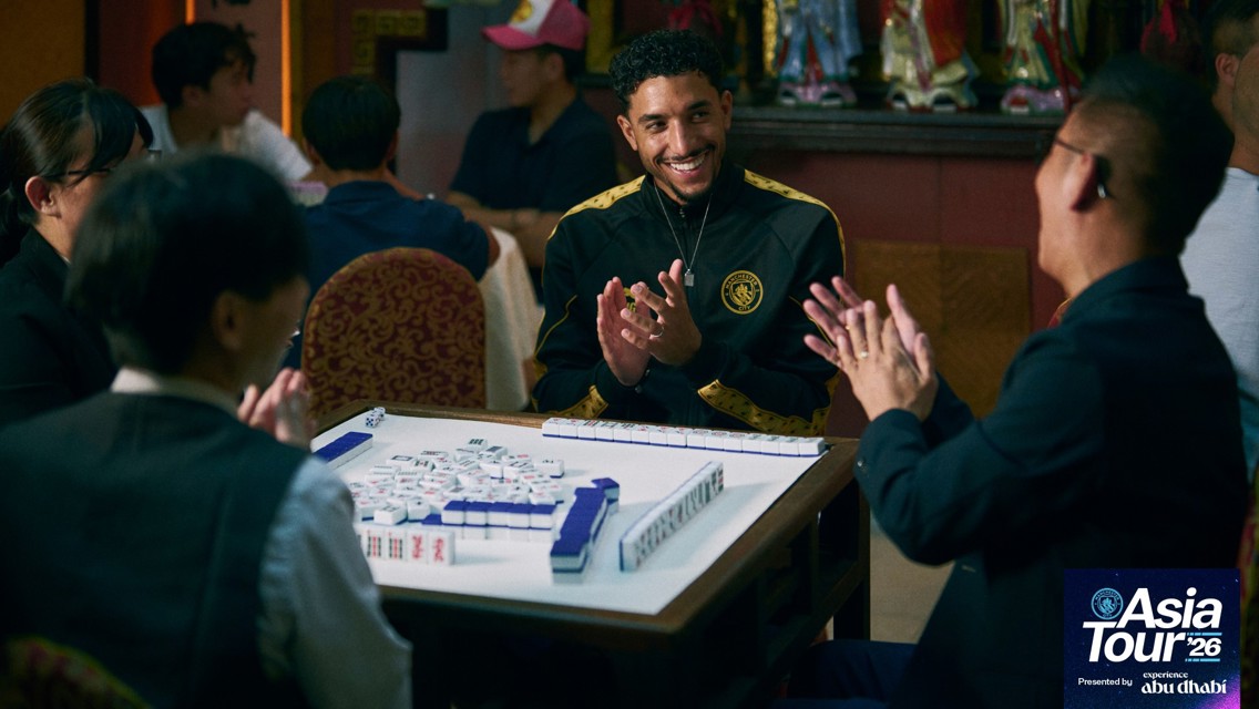 A group of people are engaged in a game of Mahjong at a table. One person in a black Manchester City jacket is smiling and clapping, surrounded by others. The event is part of 'Asia Tour '26 presented by Experience Abu Dhabi.'