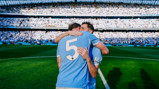 Two soccer players in blue jerseys embrace after a match on the field with a crowd in the background.