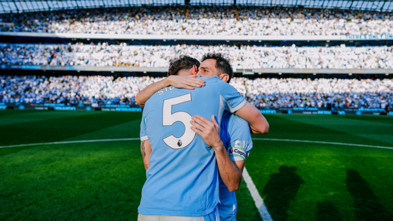 Two soccer players in blue jerseys embrace after a match on the field with a crowd in the background.