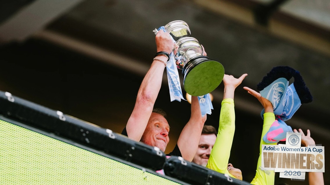 People lifting the Adobe Women's FA Cup trophy in celebration, indicating the winners of the 2026 tournament. The trophy is adorned with ribbons and scarves, and a 'Winners 2026' sign is visible.