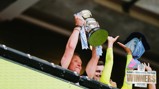People lifting the Adobe Women's FA Cup trophy in celebration, indicating the winners of the 2026 tournament. The trophy is adorned with ribbons and scarves, and a 'Winners 2026' sign is visible.