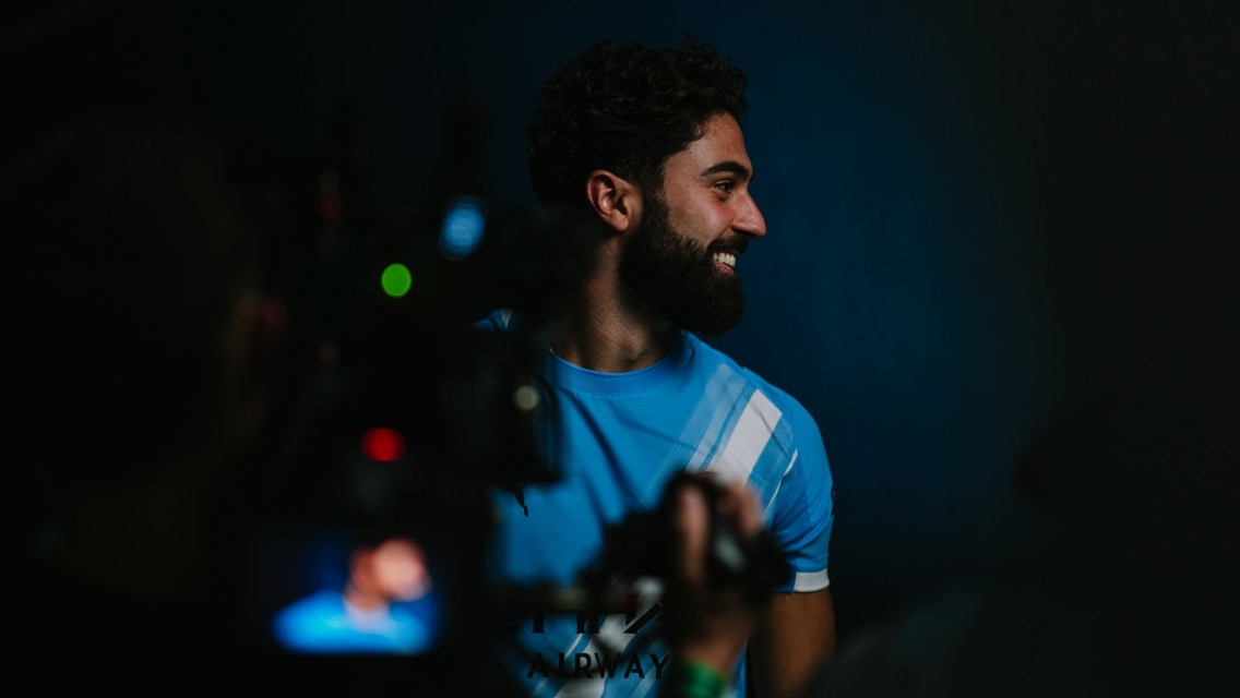 A person wearing a blue Manchester City football jersey with white stripes is being filmed in a dimly lit setting.