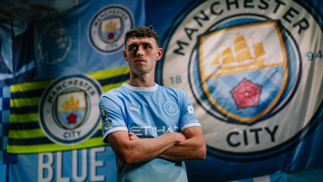 A person wearing a Manchester City football jersey stands in front of Manchester City and Premier League flags.