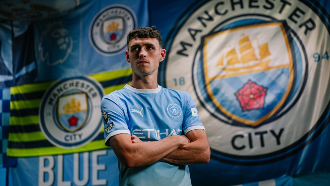A person wearing a Manchester City football jersey stands in front of Manchester City and Premier League flags.