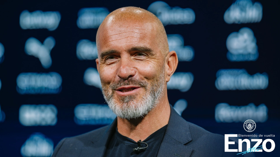 A man with a beard and bald head smiles during a press event. The background features logos including Puma and Manchester City. Text reads 'Bienvenido de vuelta Enzo'.