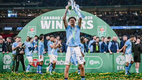 A football player celebrates with the Carabao Cup trophy in front of a 'Winners 2026' banner. Teammates and officials are in the background on a confetti-covered field.