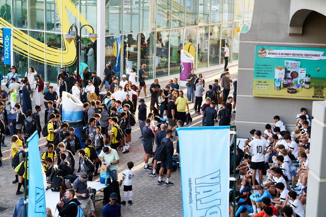 A large group of young athletes in team uniforms and backpacks gathered outside a modern building with banners and advertisements.