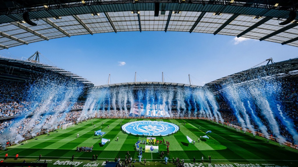 Etihad Stadium filled with fans during a Manchester City match. Blue and white confetti fills the air as large banners are displayed on the field and stands. The atmosphere is celebratory.