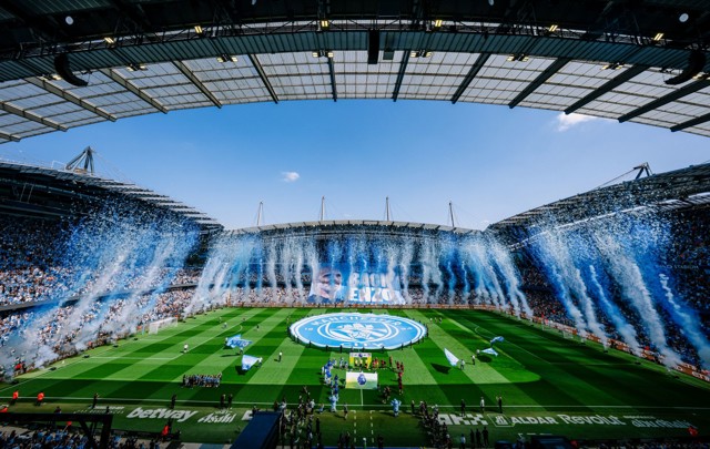 Etihad Stadium filled with fans during a Manchester City match. Blue and white confetti fills the air as large banners are displayed on the field and stands. The atmosphere is celebratory.