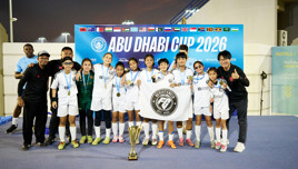 Soccer team celebrates winning the Abu Dhabi Cup 2026. Players in white uniforms hold medals and a team banner, standing behind a large trophy. International flags are displayed on the backdrop.