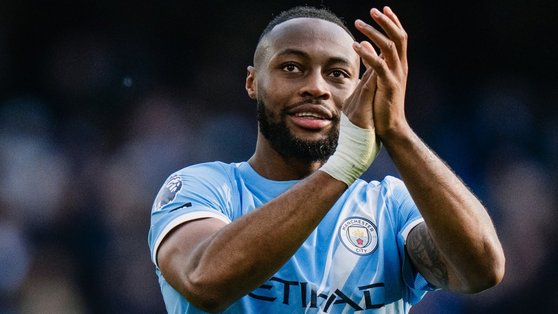 A Manchester City player wearing a blue jersey clapping on the field with a blurred face. The Premier League logo is visible on the sleeve.