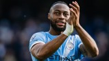 A Manchester City player wearing a blue jersey clapping on the field with a blurred face. The Premier League logo is visible on the sleeve.