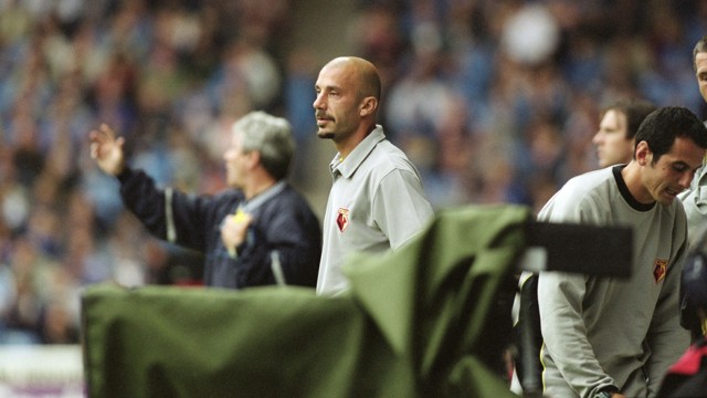 A football coach in a gray Watford FC shirt stands on the sideline during a game. Another person, possibly an assistant, is beside him. The background shows a blurred audience.