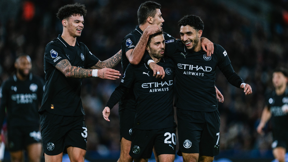 Football players in black Manchester City kits celebrating after a goal on the field.