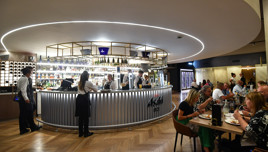 A modern bar with a curved counter, illuminated shelves, and wine bottles. Staff interact with guests. Nearby tables have diners enjoying meals. Asahi branding is visible on the counter.