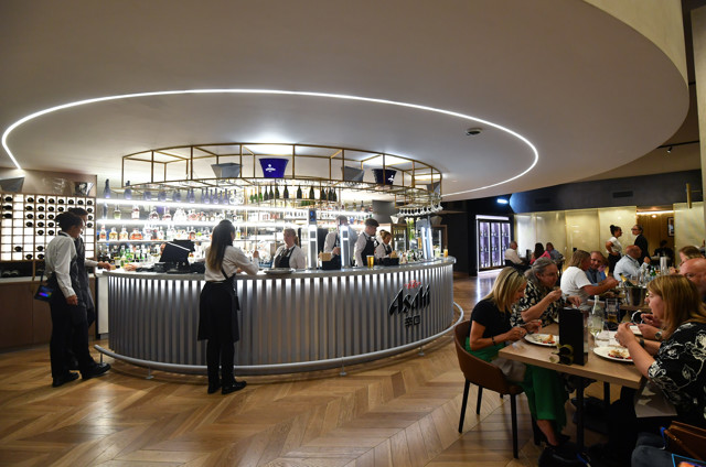 A modern bar with a curved counter, illuminated shelves, and wine bottles. Staff interact with guests. Nearby tables have diners enjoying meals. Asahi branding is visible on the counter.