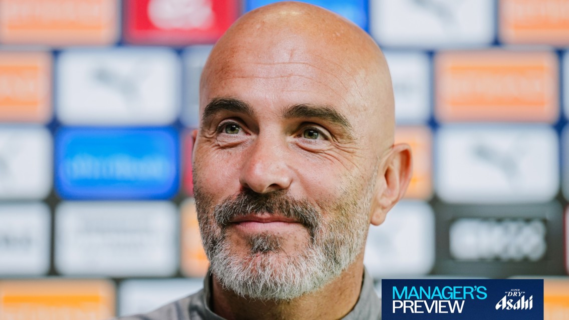 A bald man with a beard smiling during a press conference. Background features a wall with blurred logos.