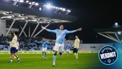 A Manchester City player celebrates a goal with outstretched arms during a match at night, wearing the team's blue kit with Etihad Airways sponsorship. Other players and stadium lights visible in the background.