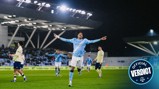 A Manchester City player celebrates a goal with outstretched arms during a match at night, wearing the team's blue kit with Etihad Airways sponsorship. Other players and stadium lights visible in the background.