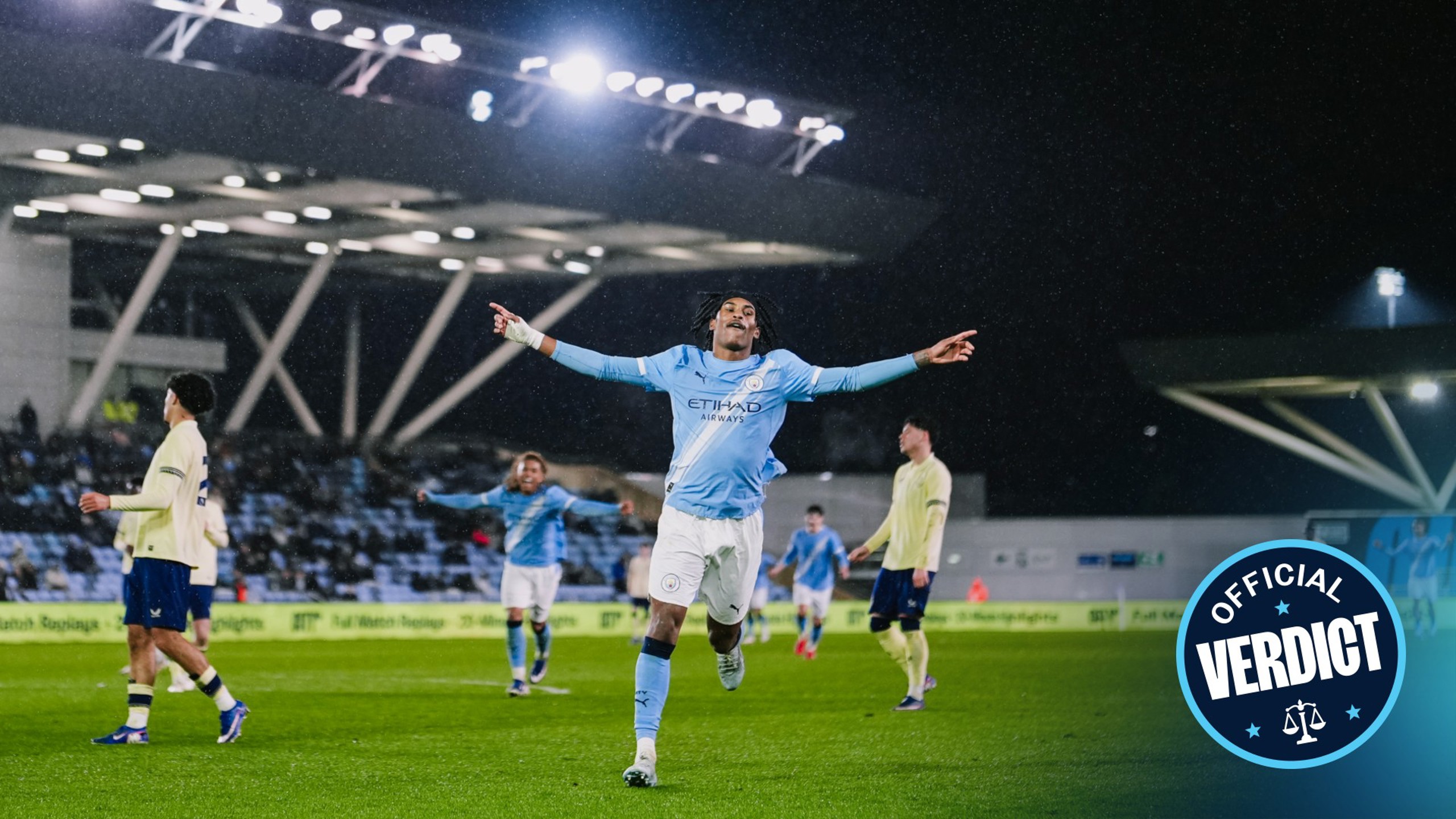 A Manchester City player celebrates a goal with outstretched arms during a match at night, wearing the team's blue kit with Etihad Airways sponsorship. Other players and stadium lights visible in the background.