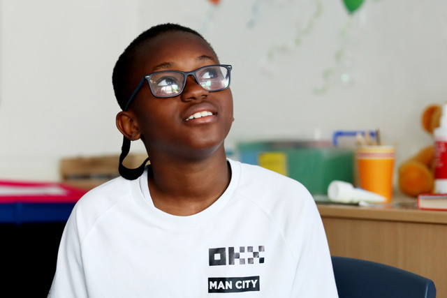 Boy wearing a Manchester City white sweatshirt featuring the OKX logo, in a room with blurred background including colorful objects and table.