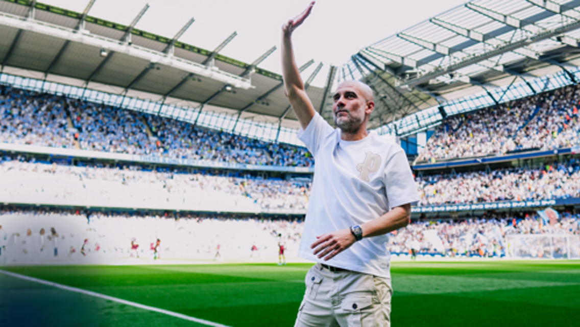 A person raises an arm in celebration at a football stadium, with a large crowd in the background.