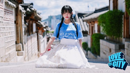 A woman wearing a light blue sports jersey and a white skirt with gold detailing, standing in an alley of Bukchon Hanok Village, Seoul. The image has a 'Love This City' logo at the bottom right corner.