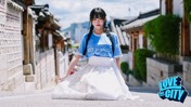 A woman wearing a light blue sports jersey and a white skirt with gold detailing, standing in an alley of Bukchon Hanok Village, Seoul. The image has a 'Love This City' logo at the bottom right corner.