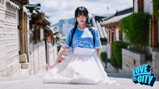 A woman wearing a light blue sports jersey and a white skirt with gold detailing, standing in an alley of Bukchon Hanok Village, Seoul. The image has a 'Love This City' logo at the bottom right corner.