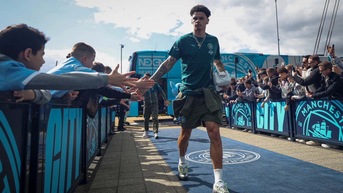 A Manchester City player wearing a green training outfit greets fans as he walks past barriers with Manchester City logos.
